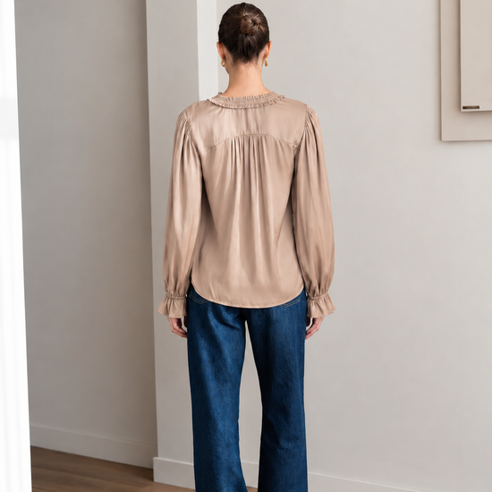 A woman viewed from behind wearing a beige long-sleeve blouse with gathered detailing and dark blue wide-leg jeans, standing in a minimalist room with light gray walls and a wood floor.