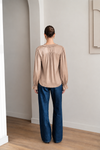 A woman viewed from behind wearing a beige long-sleeve blouse with gathered detailing and dark blue wide-leg jeans, standing in a minimalist room with light gray walls and a wood floor.