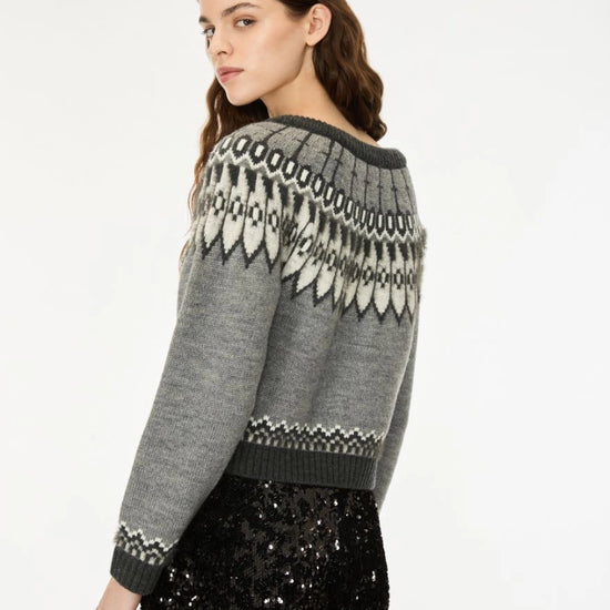 A woman wearing a gray knit sweater with a black and white Fair Isle pattern across the shoulders and sleeves, paired with a black sequined skirt, photographed against a plain white studio background.
