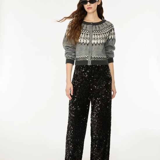 A model wearing a gray and white patterned knit cardigan with black trim, paired with black sequined wide-leg pants, sunglasses, and black shoes, photographed against a plain white studio background.