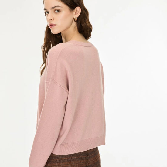 A woman with long wavy brown hair wearing a dusty pink long-sleeve knit sweater and brown patterned trousers, standing against a plain white studio background.