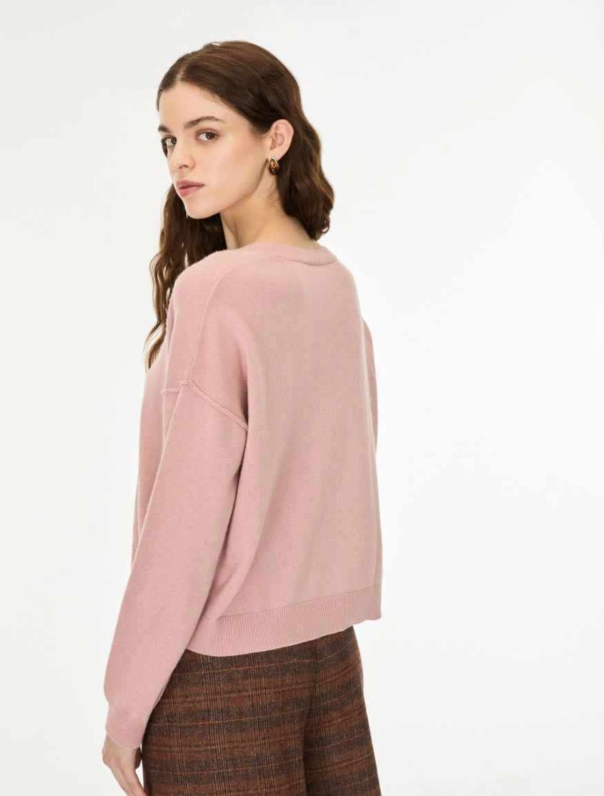 A woman with long wavy brown hair wearing a dusty pink long-sleeve knit sweater and brown patterned trousers, standing against a plain white studio background.