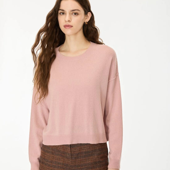 A woman with long wavy brown hair wearing a dusty pink long-sleeve knit sweater and brown patterned trousers, standing against a plain white studio background.