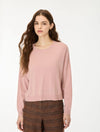 A woman with long wavy brown hair wearing a dusty pink long-sleeve knit sweater and brown patterned trousers, standing against a plain white studio background.