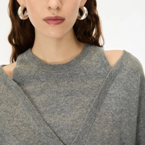A close-up of a woman wearing a gray knit sweater with a high crew neck and shoulder cutouts, paired with chunky silver hoop earrings against a plain white background.