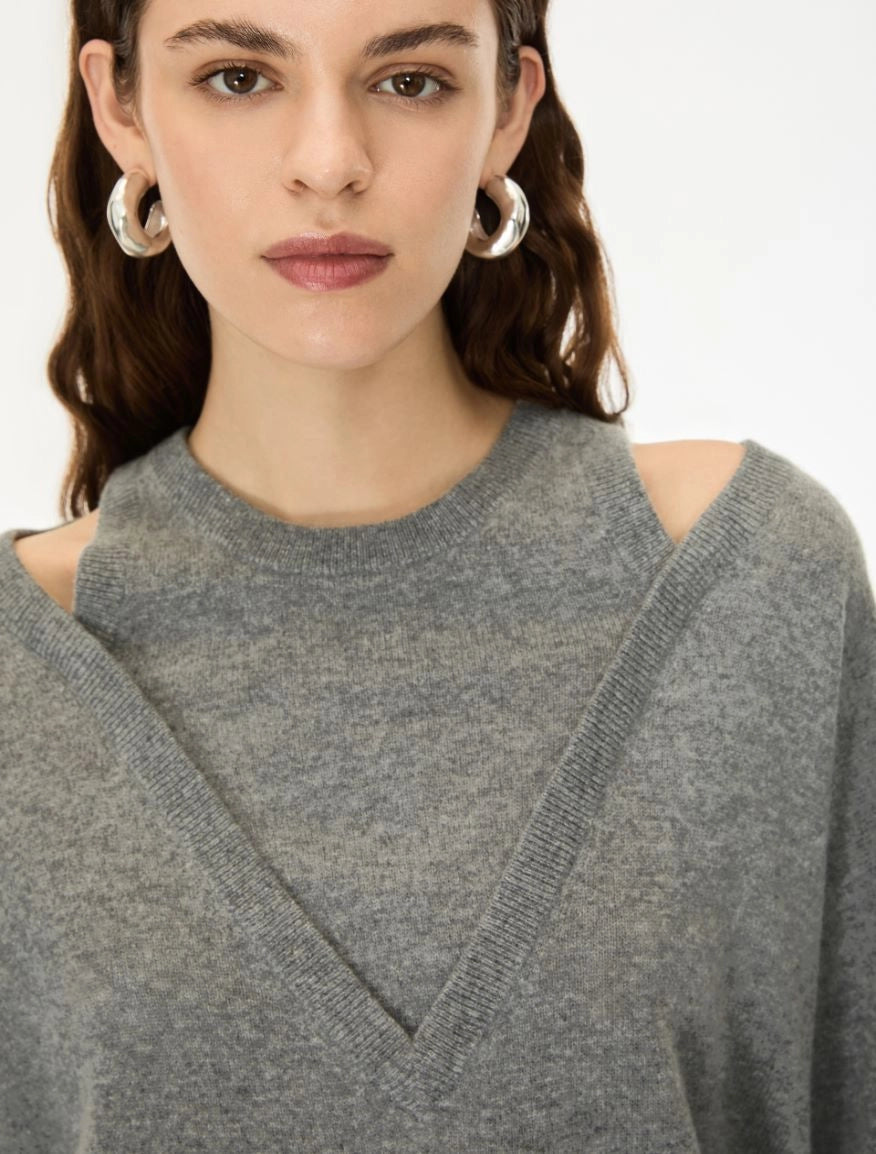 A close-up of a woman wearing a gray knit sweater with a high crew neck and shoulder cutouts, paired with chunky silver hoop earrings against a plain white background.