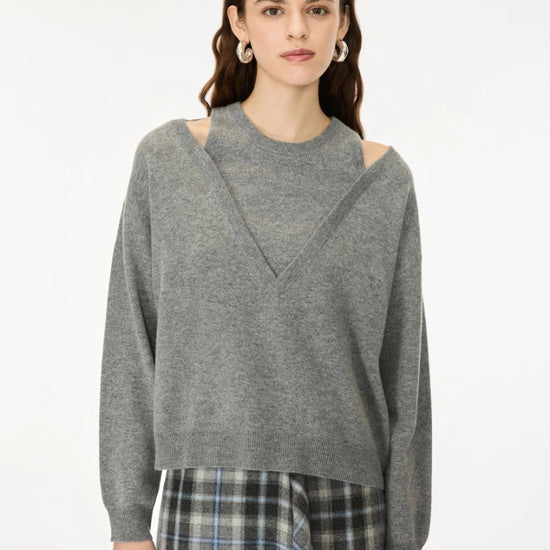 A woman wearing a heather gray knit sweater with shoulder cutouts and a V-shaped neckline, paired with a plaid skirt, against a plain white studio background.