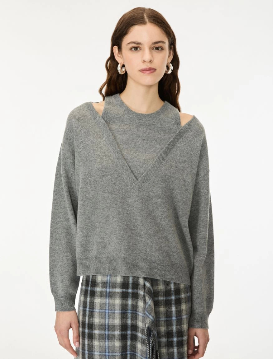 A woman wearing a heather gray knit sweater with shoulder cutouts and a V-shaped neckline, paired with a plaid skirt, against a plain white studio background.