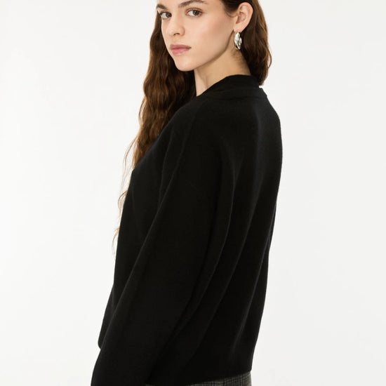 A woman with long brown hair is shown from a side-back angle wearing a black knit sweater and gray plaid trousers against a plain white studio background.