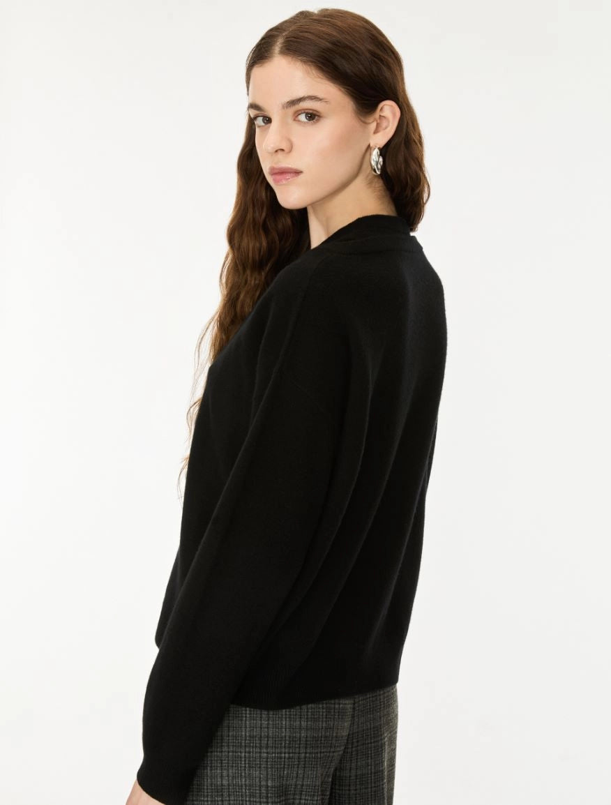 A woman with long brown hair is shown from a side-back angle wearing a black knit sweater and gray plaid trousers against a plain white studio background.