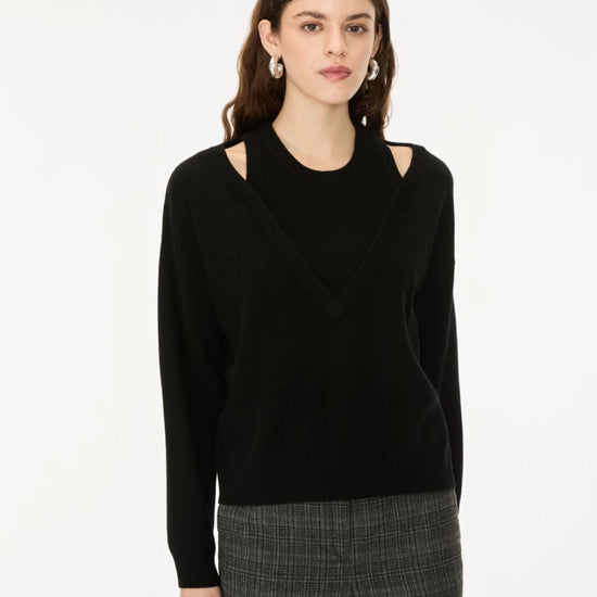 A woman modeling a black knit sweater with shoulder cutouts and a V-neckline, paired with gray plaid trousers, against a plain white studio background.
