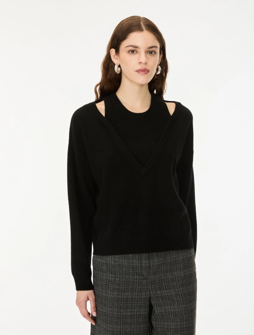 A woman modeling a black knit sweater with shoulder cutouts and a V-neckline, paired with gray plaid trousers, against a plain white studio background.