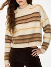 A woman wearing a fuzzy knit sweater with horizontal stripes in cream, tan, brown, and mustard yellow, paired with dark brown pants against a plain white background.