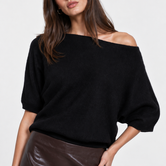 A model wearing a black off-the-shoulder knit top, a shiny dark brown faux-leather midi skirt, and black knee-high boots, posed against a light gray studio background.