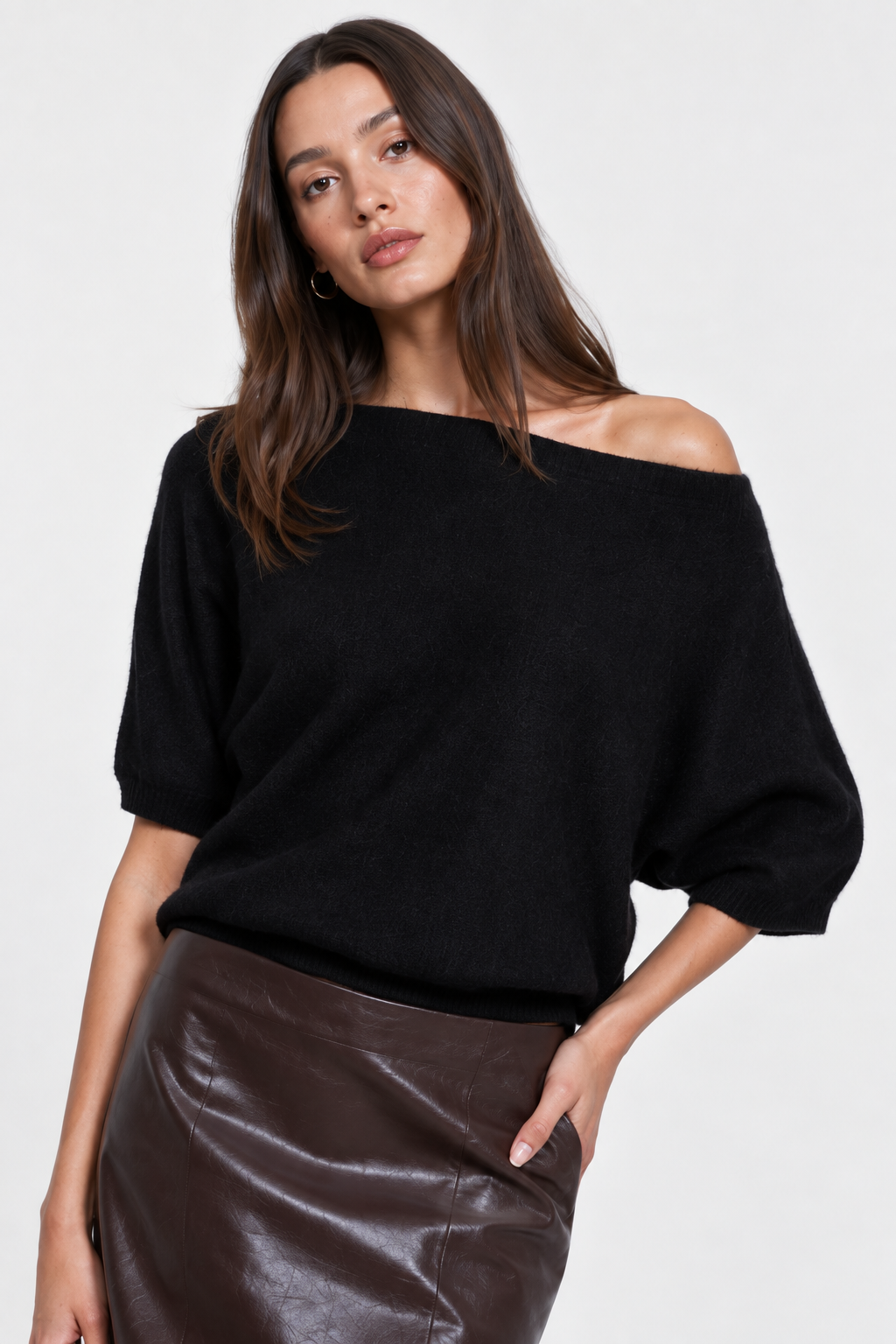 A model wearing a black off-the-shoulder knit top, a shiny dark brown faux-leather midi skirt, and black knee-high boots, posed against a light gray studio background.