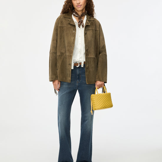 Woman wearing a green jacket, white shirt, blue jeans, and brown shoes, holding a yellow handbag on a white background.
