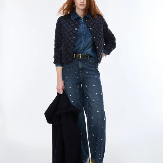 A model stands against a plain white studio background wearing a dark quilted jacket over a blue denim shirt and dark blue wide-leg jeans with small white embroidered details, holding a folded black coat.