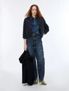 A model stands against a plain white studio background wearing a dark quilted jacket over a blue denim shirt and dark blue wide-leg jeans with small white embroidered details, holding a folded black coat.