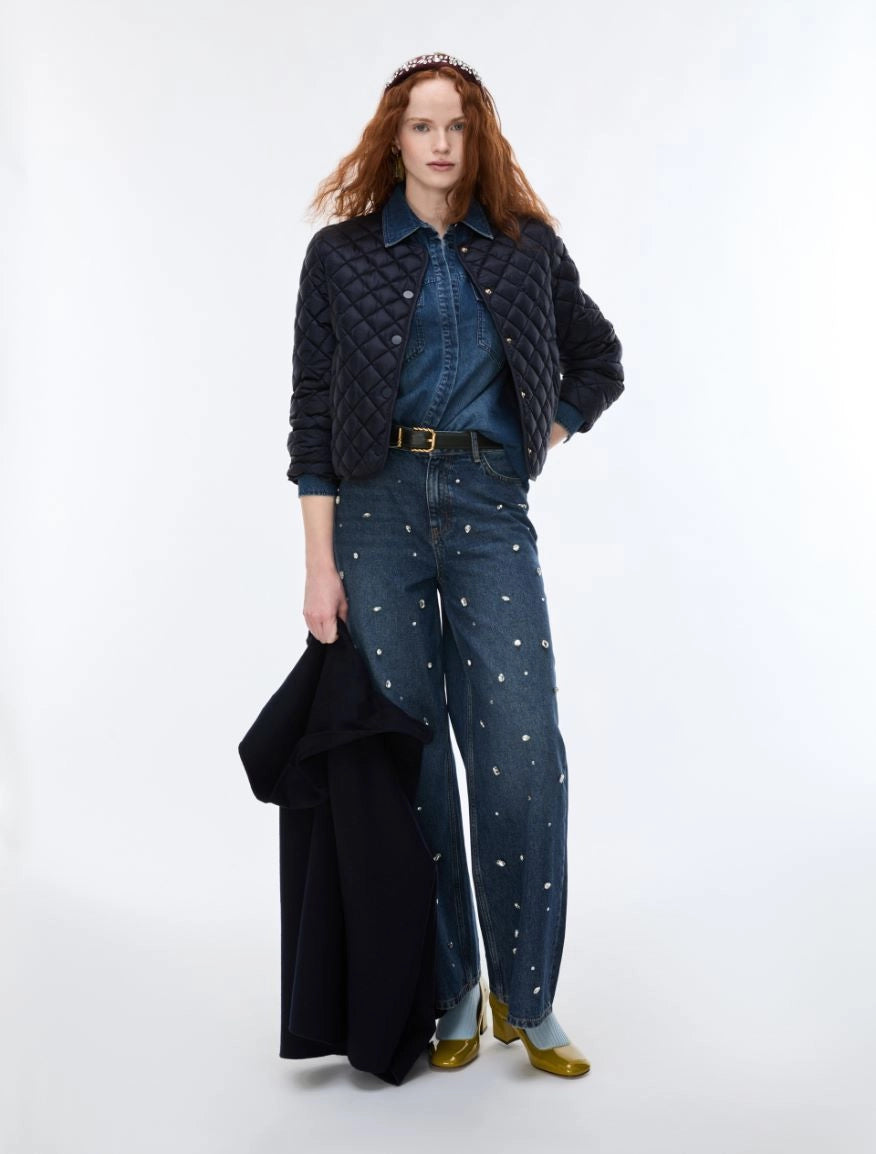 A model stands against a plain white studio background wearing a dark quilted jacket over a blue denim shirt and dark blue wide-leg jeans with small white embroidered details, holding a folded black coat.