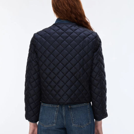 A woman viewed from the back wearing a dark navy quilted jacket and blue jeans against a plain white studio background.