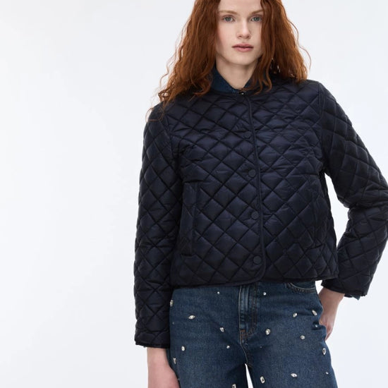 A model with red hair wearing a dark navy blue diamond-quilted cropped jacket and blue jeans with silver studs, standing against a plain white studio background.