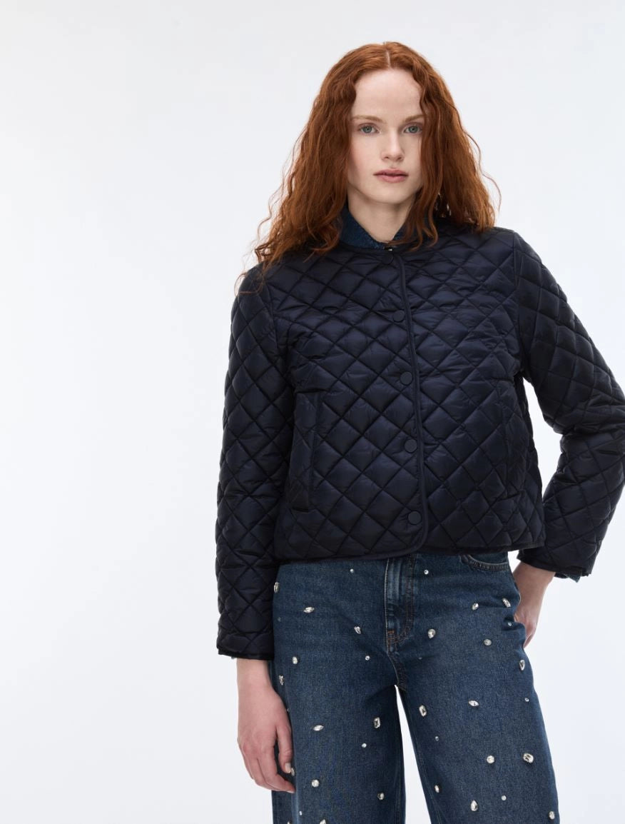 A model with red hair wearing a dark navy blue diamond-quilted cropped jacket and blue jeans with silver studs, standing against a plain white studio background.