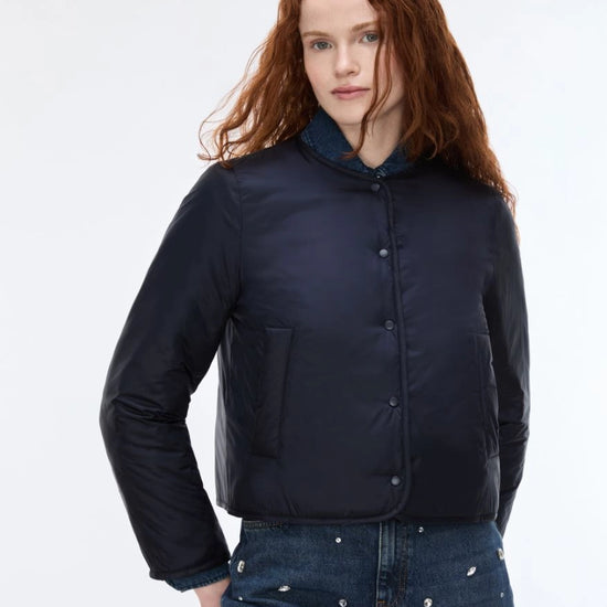 A model with red hair wearing a dark navy blue cropped puffer jacket with a stand-up collar and front buttons, paired with blue jeans with silver studs, against a plain white studio background.