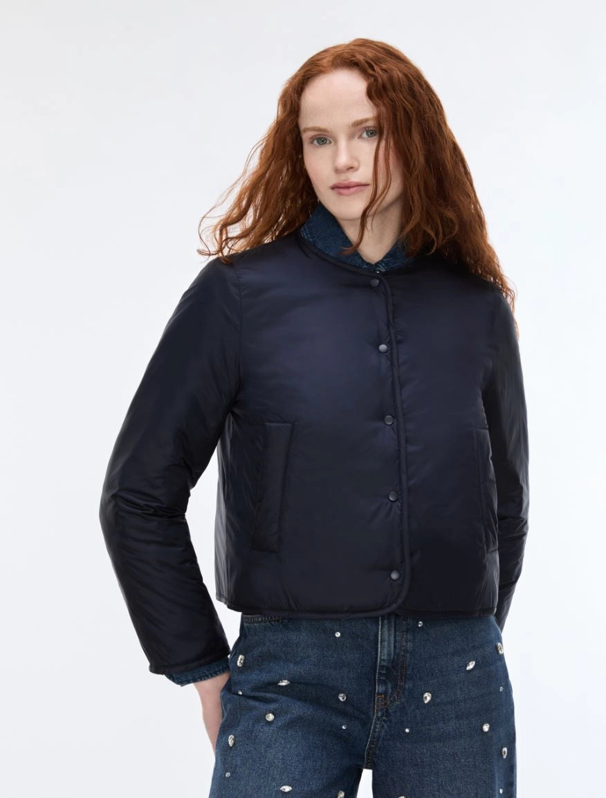 A model with red hair wearing a dark navy blue cropped puffer jacket with a stand-up collar and front buttons, paired with blue jeans with silver studs, against a plain white studio background.