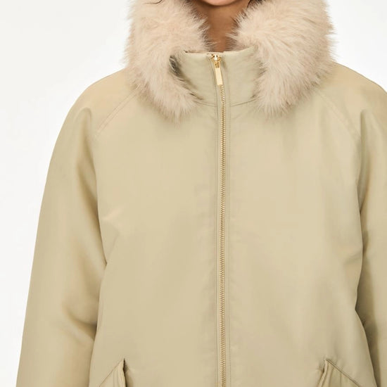 A close-up of a person wearing a beige winter parka with a fluffy cream faux-fur collar and a front zipper, against a plain light background.