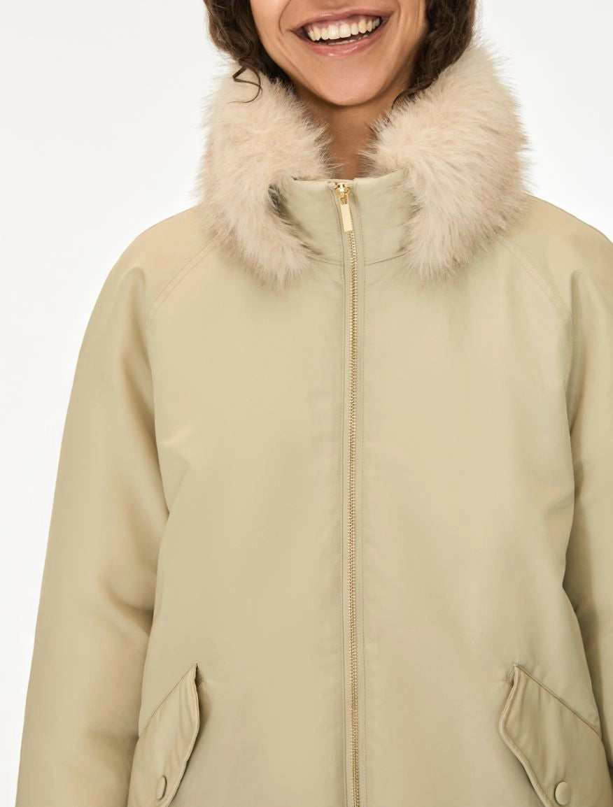 A close-up of a person wearing a beige winter parka with a fluffy cream faux-fur collar and a front zipper, against a plain light background.
