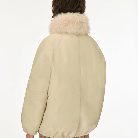 A person shown from the back wearing a cream-colored puffer jacket with a fluffy light beige faux-fur collar, paired with brown and beige checkered trousers, against a plain white studio background.