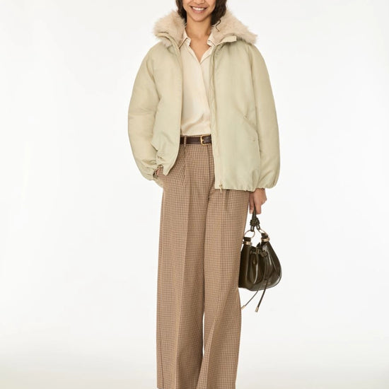 A woman stands against a plain white studio background wearing a light beige puffer jacket with a fluffy collar, a cream blouse, wide-leg tan houndstooth trousers, dark brown shoes, and holding a dark green handbag.