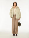 A woman stands against a plain white studio background wearing a light beige puffer jacket with a fluffy collar, a cream blouse, wide-leg tan houndstooth trousers, dark brown shoes, and holding a dark green handbag.