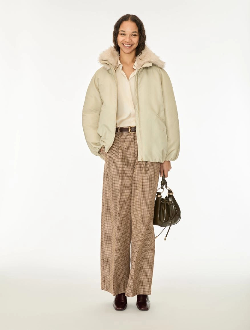 A woman stands against a plain white studio background wearing a light beige puffer jacket with a fluffy collar, a cream blouse, wide-leg tan houndstooth trousers, dark brown shoes, and holding a dark green handbag.