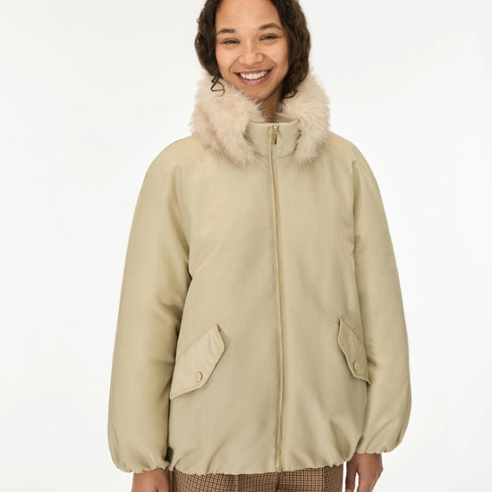 A smiling woman wearing a beige puffer jacket with a fluffy cream faux-fur collar, paired with brown plaid trousers, against a plain white studio background.