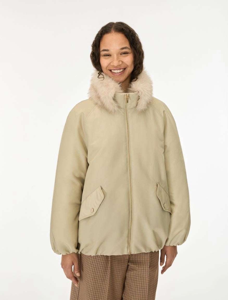 A smiling woman wearing a beige puffer jacket with a fluffy cream faux-fur collar, paired with brown plaid trousers, against a plain white studio background.