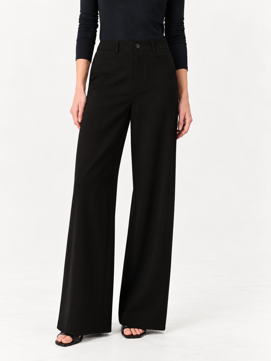 A model wearing black wide-leg trousers and a black long-sleeve top, standing against a plain white studio background.