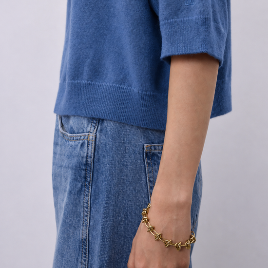 A close-up of a person wearing a blue knit short-sleeve top and blue jeans, with a gold chain bracelet on the wrist and a gold ring on the finger, against a plain light gray background.
