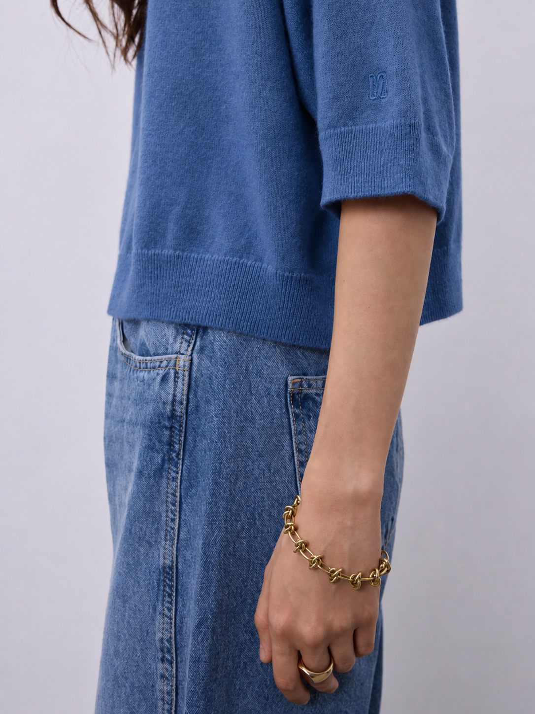 A close-up of a person wearing a blue knit short-sleeve top and blue jeans, with a gold chain bracelet on the wrist and a gold ring on the finger, against a plain light gray background.