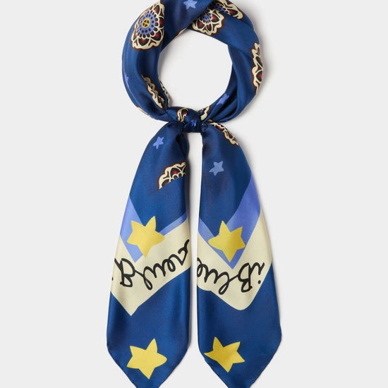 A blue silk scarf with a celestial print featuring yellow stars, a cream banner with black cursive text, and gold-toned ornamental details, shown on a light gray background.