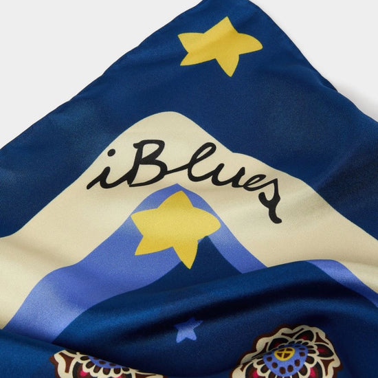 A close-up of a blue scarf featuring a mountain design with yellow stars, the script text 'iBlues', and ornate floral motifs.