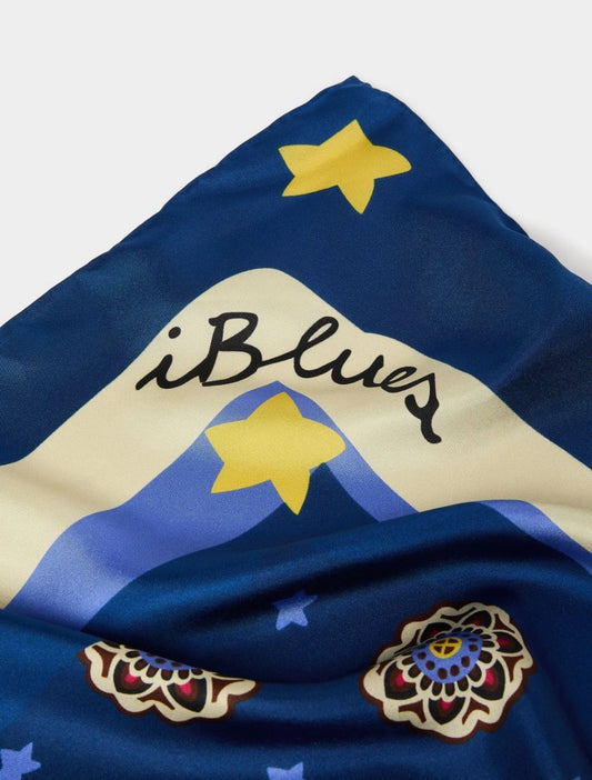A close-up of a blue scarf featuring a mountain design with yellow stars, the script text 'iBlues', and ornate floral motifs.