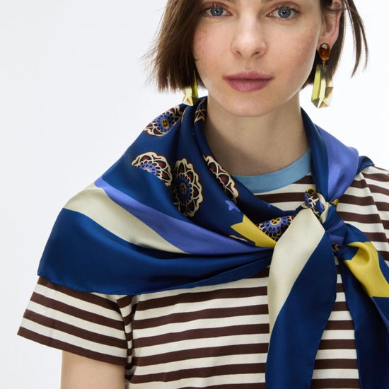 A woman wearing a blue silk scarf with cream and yellow stripes and floral motifs, styled around her neck over a brown and cream striped t-shirt against a plain white background.