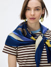 A woman wearing a blue silk scarf with cream and yellow stripes and floral motifs, styled around her neck over a brown and cream striped t-shirt against a plain white background.