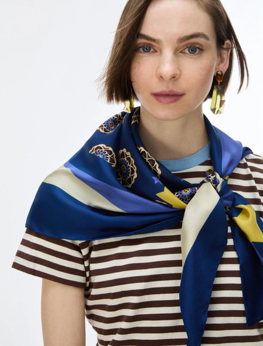 A woman wearing a blue silk scarf with cream and yellow stripes and floral motifs, styled around her neck over a brown and cream striped t-shirt against a plain white background.