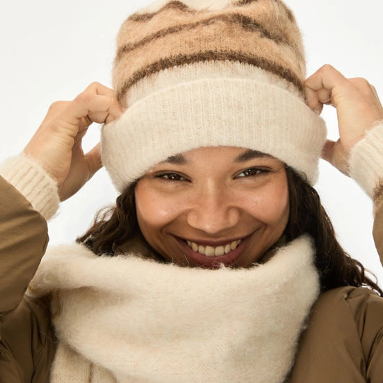 A smiling woman wearing a cream knit beanie with brown stripes and a matching cream knit neck warmer, adjusting the beanie with both hands against a plain white background.