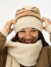 A smiling woman wearing a cream knit beanie with brown stripes and a matching cream knit neck warmer, adjusting the beanie with both hands against a plain white background.