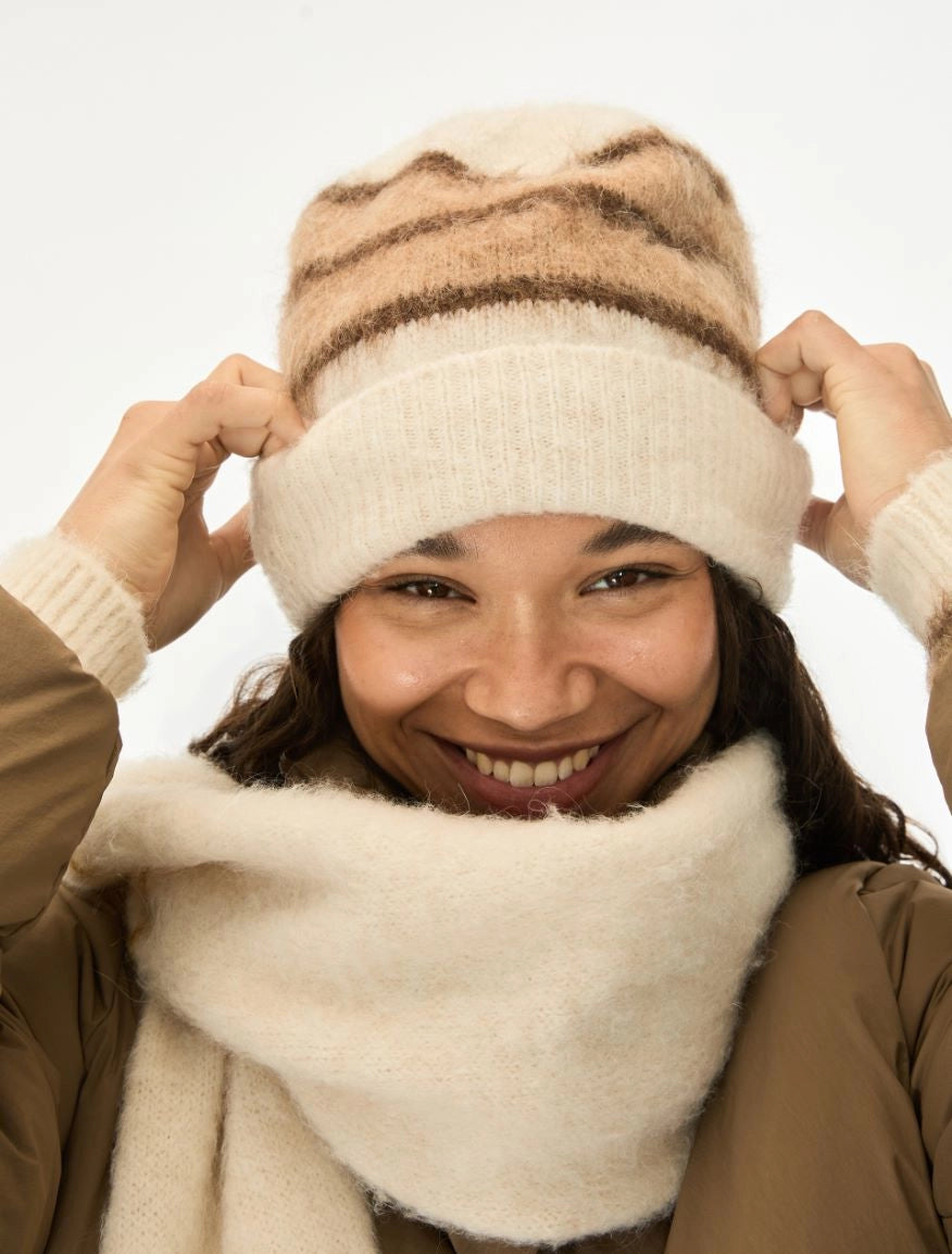 A smiling woman wearing a cream knit beanie with brown stripes and a matching cream knit neck warmer, adjusting the beanie with both hands against a plain white background.