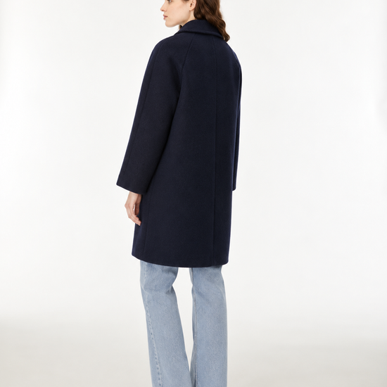A woman modeling a dark navy blue wool-blend coat with front buttons, paired with light blue jeans and brown shoes, standing against a plain white studio background.