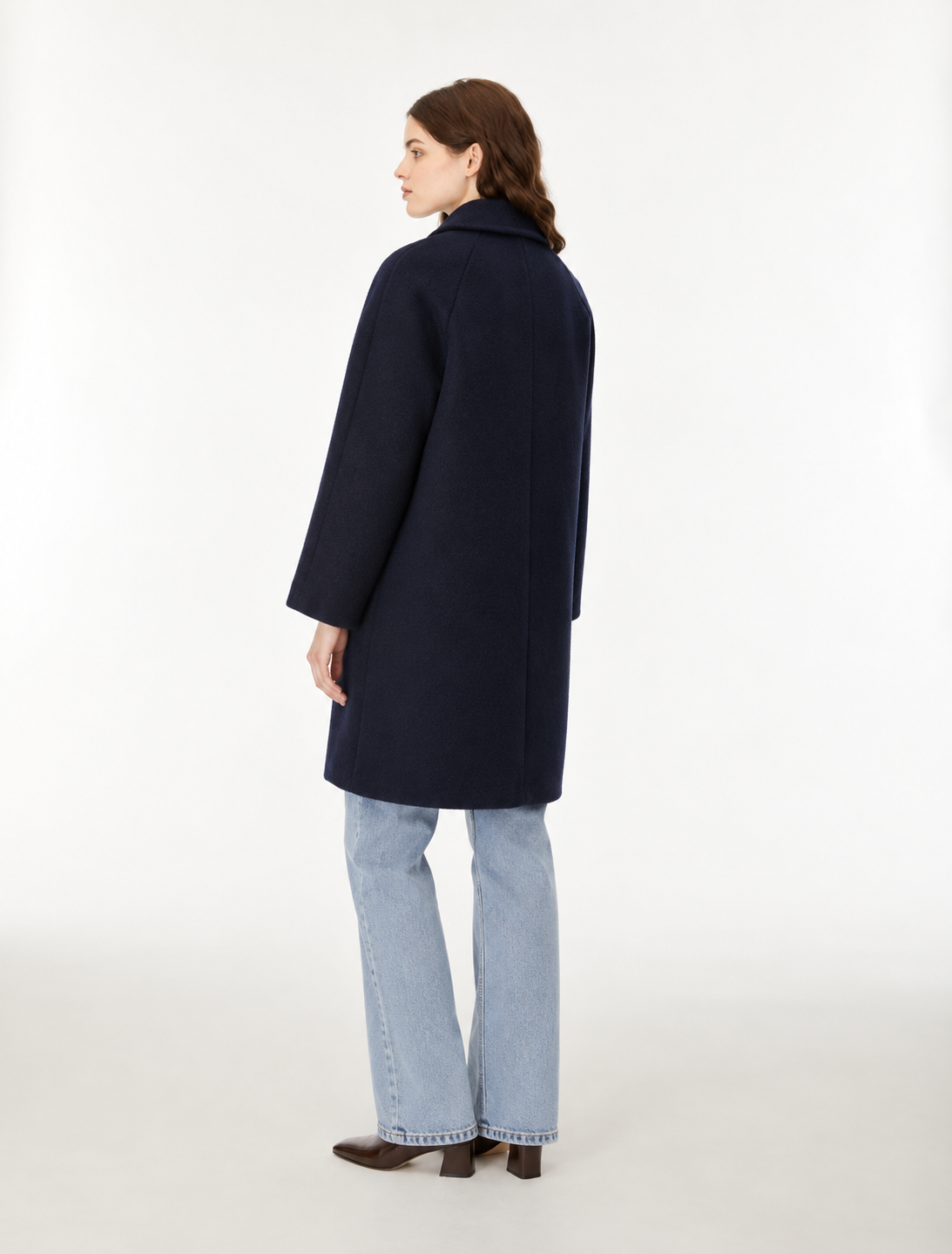 A woman modeling a dark navy blue wool-blend coat with front buttons, paired with light blue jeans and brown shoes, standing against a plain white studio background.